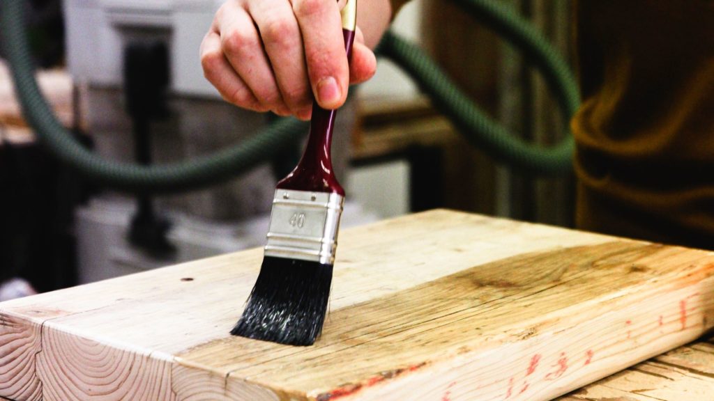 Someone painting varnish onto a piece of wood with a dark red paint brush.
